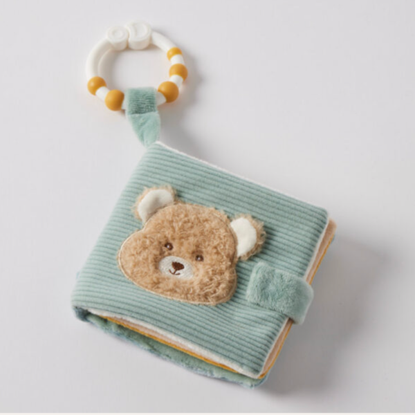 A baby soft toy book in sage green corduroy fabric with a plush teddy bear face on the cover, featuring a matching green strap closure and an attached white and yellow beaded ring rattle, photographed on a light gray background.