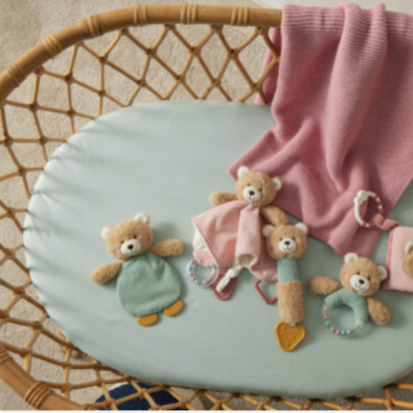 A styled nursery scene showing a round pale blue mattress or cushion inside a woven rattan bassinet, with soft teddy bear toys and a pink knit blanket arranged on top.